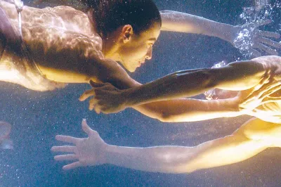 Unterwasserszene aus Dido und Aeneas: Zwei Darstellende schweben einander gegenüber in einem Wasserbecken, ihre Körper sanft im Raum gehalten, die Arme einander entgegenstreckend, während Luftblasen zwischen ihnen aufsteigen und warmes, goldenes Licht die Szene durchzieht.